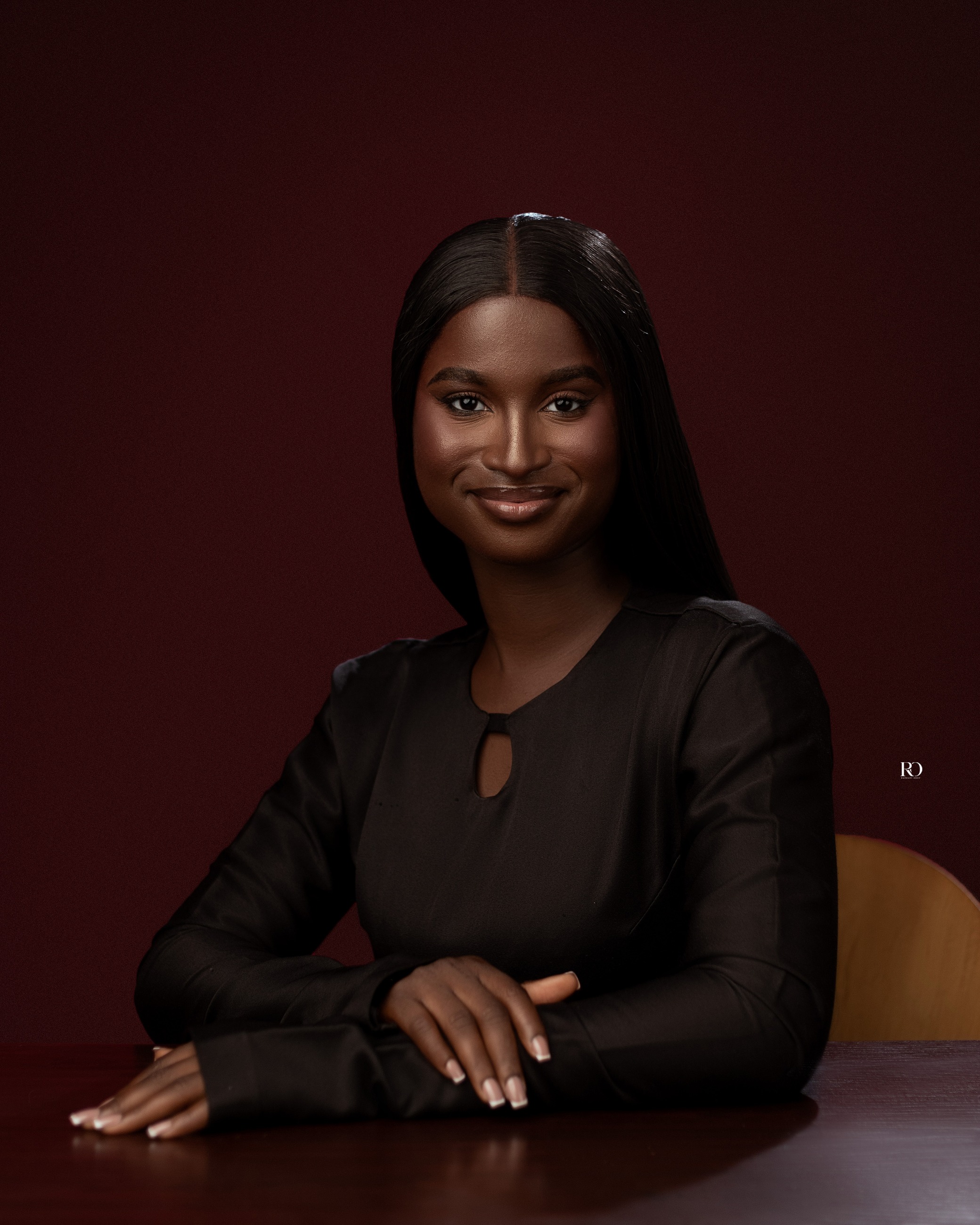 Gladys Ogo seated in a dark dress against a deep burgundy studio backdrop